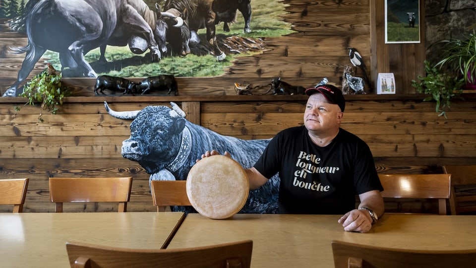 Mann sitzt in Chalet an einem Tisch, hält einen Laib Käse.