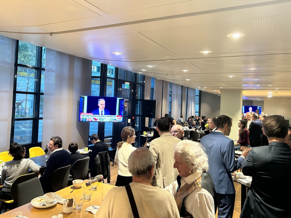 Menschenmenge in einem Konferenzraum mit Fernseher an der Wand.