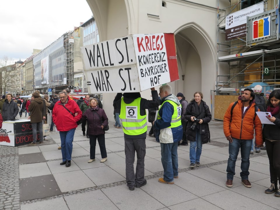 Ein Demonstrant hält ein Transparent mit den Aufschriften «Wall St. = War St.» und «Kriegskonferenz Bayrischer Hof» in die Höhe.