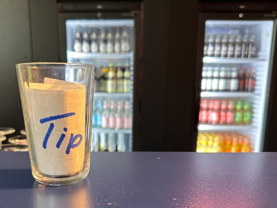 Trinkgeldglas auf Theke mit Kühlschränken im Hintergrund.