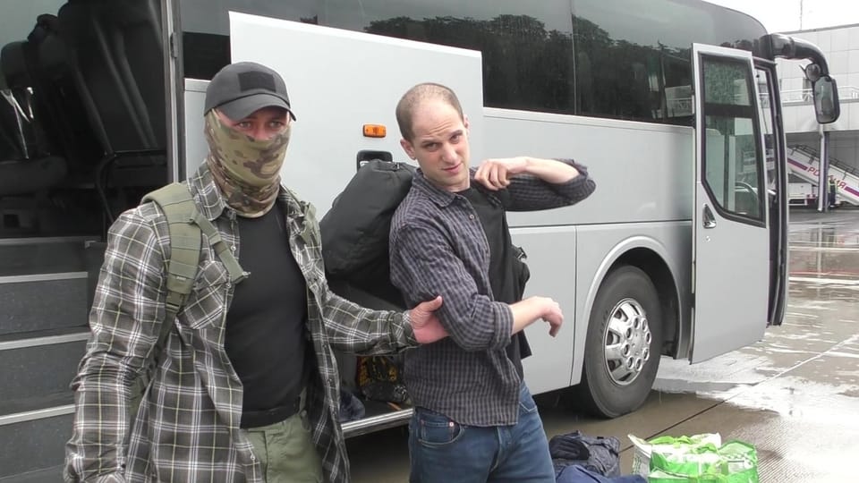Zwei Männer vor einem Reisebus, einer maskiert, der andere trägt eine Tasche.