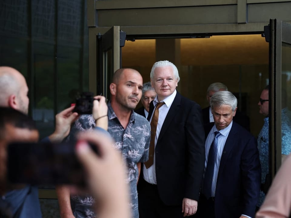Assange vor der Tür in Anzug, links verschwommen Mann mit Handy, macht Foto