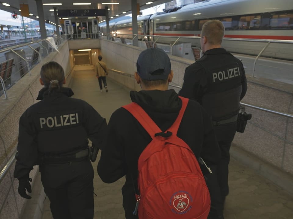 Polizisten begleiten eine Person auf dem Bahnsteig neben einem Zug.