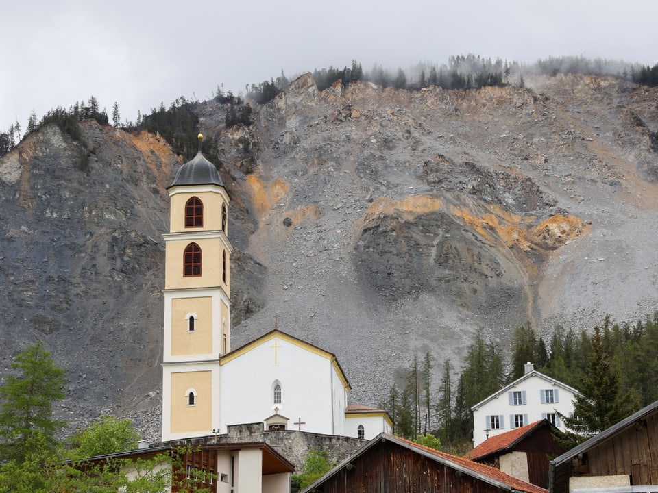 Kirchturm vor Berghang mit Geröll und Wolken.