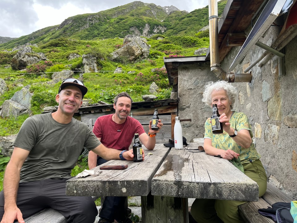 Drei Menschen geniessen Getränke an einem rustikalen Holztisch in einer Berglandschaft.