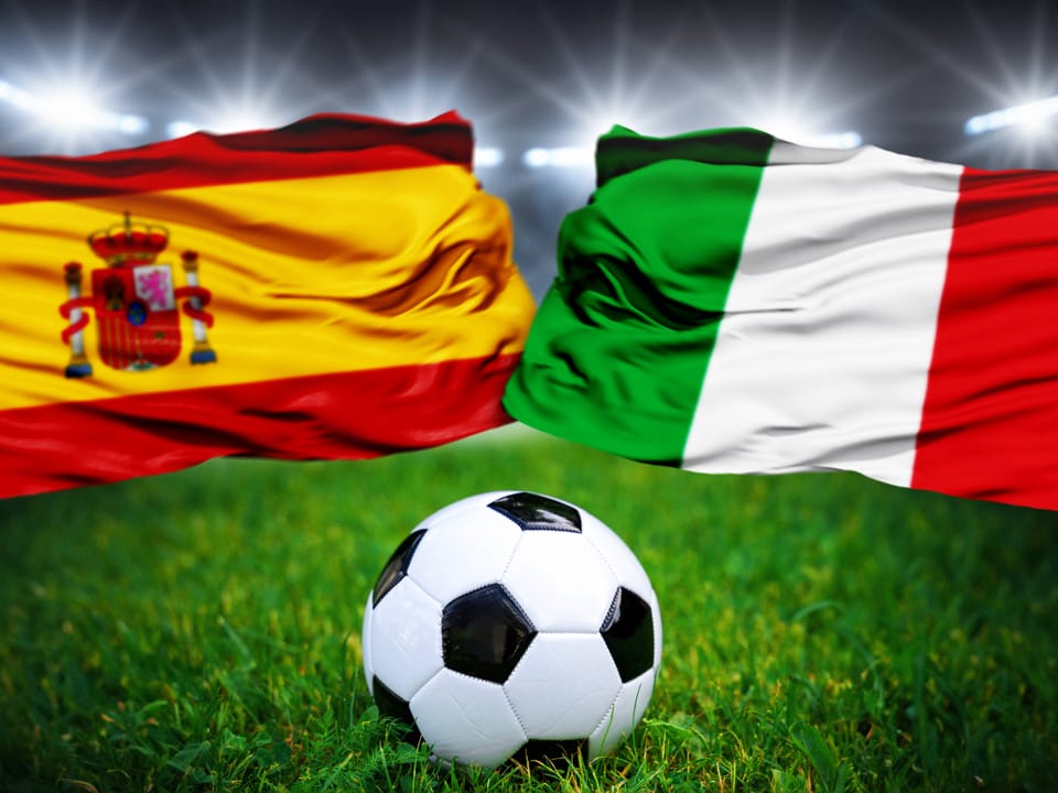 Fussball vor spanischer und italienischer Flagge auf Grasfeld.