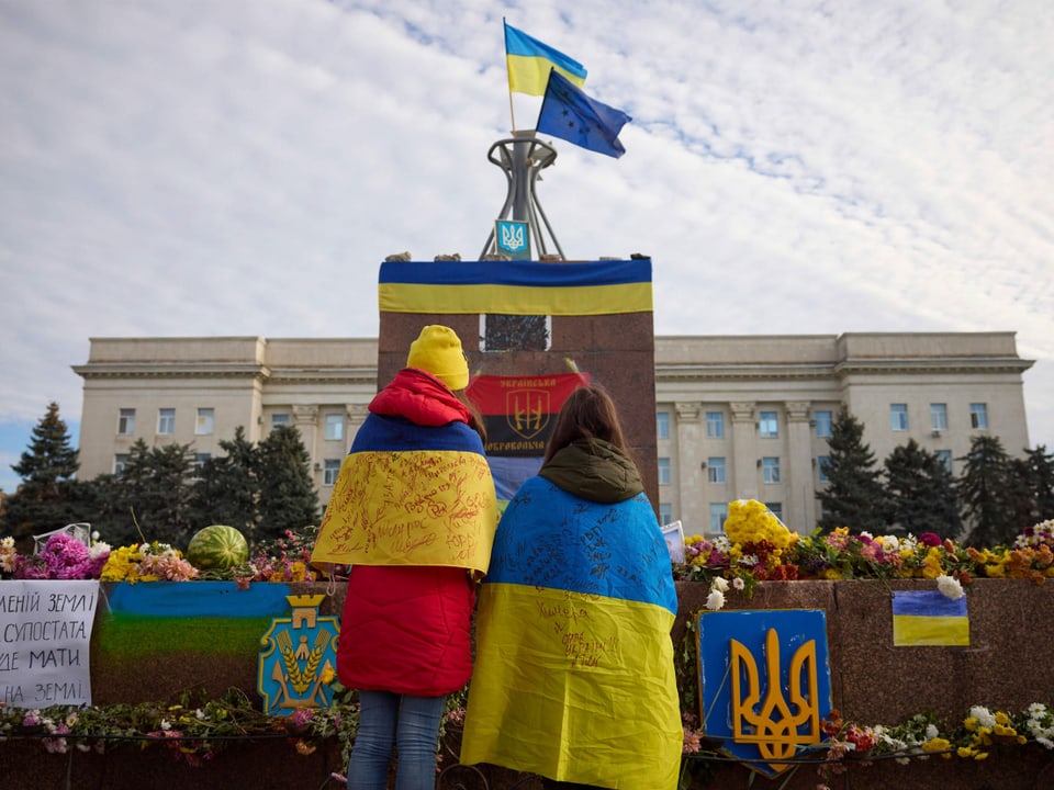 Zwei Frauen mit Ukraine-Flagge stehen vor Blumen auf einem Platz.