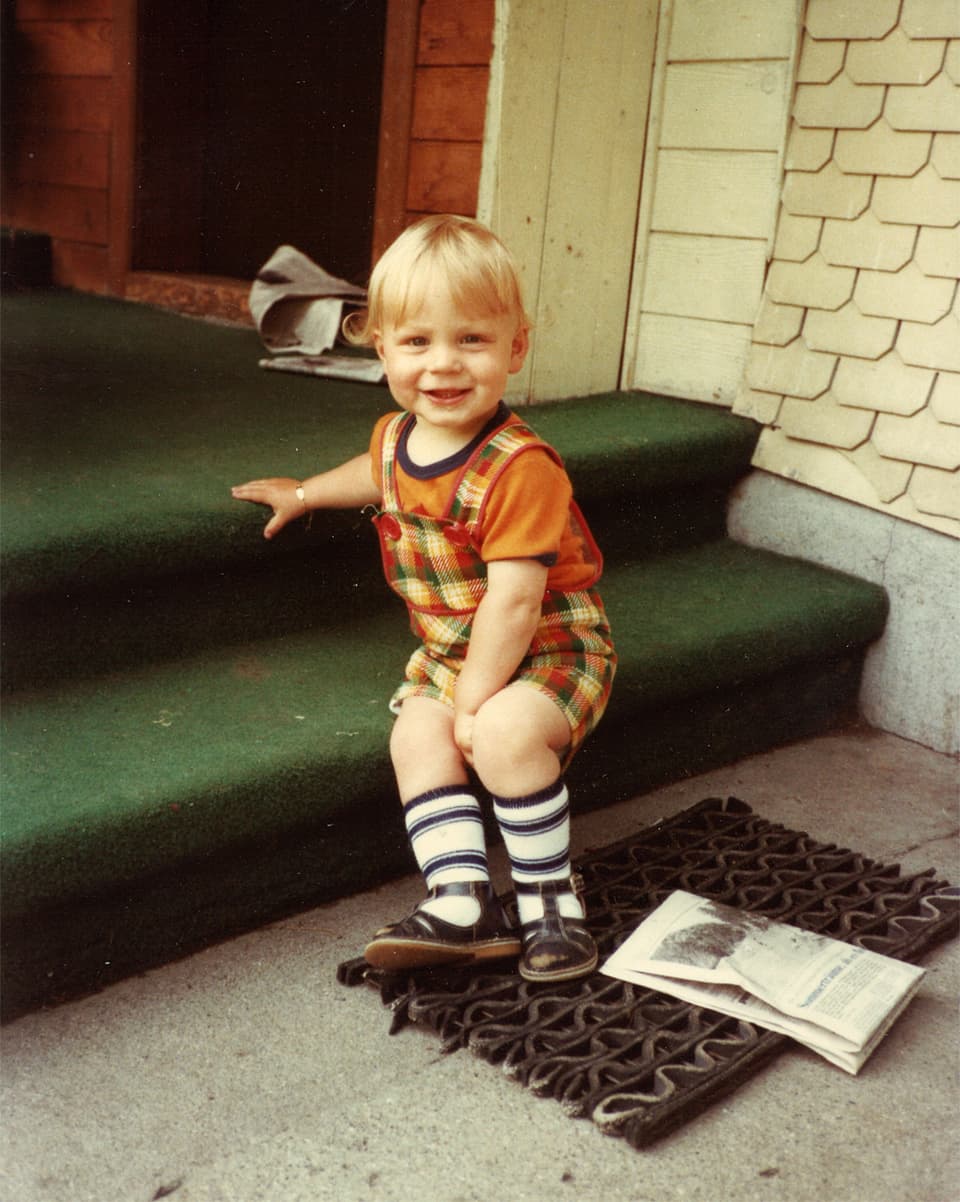 Ein kleiner Bub mit orangem T-Shirt und kurzer karierter Hose sitzt auf der Stufe einer grünen Treppe.