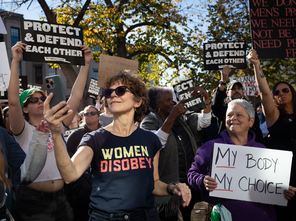 Menschenmenge bei Protest mit Schildern, Frau mit 'Women Disobey'-Shirt.