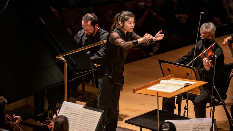 junge asiatische Frau in schwarz auf Bühne, dirigiert mit ausgestreckten Händen sitzende Musiker.