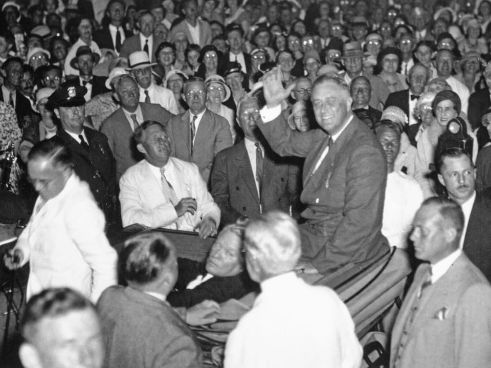 Menschenmenge bei einer Veranstaltung; im Vordergrund ein winkender Mann im Anzug auf einem Stuhl.