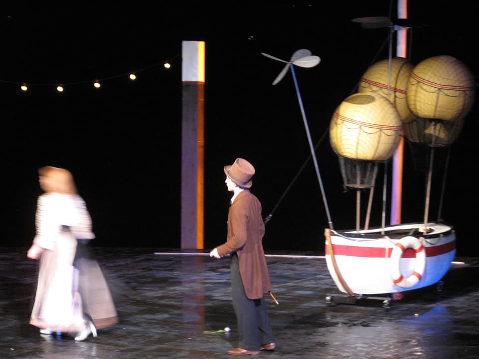 Eine Freilicht-Theaterbühne bei Nacht. Darauf ein Luftschiff, ein Mann mit Hut und Stock und eine Frau mit Rock.
