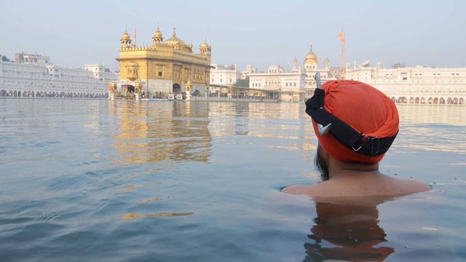 Ein junge mit Turban beim Bad schleudert mit seinen Händen Wasser in die Luft. Im Hintergrund der Tempel von Amritsar 