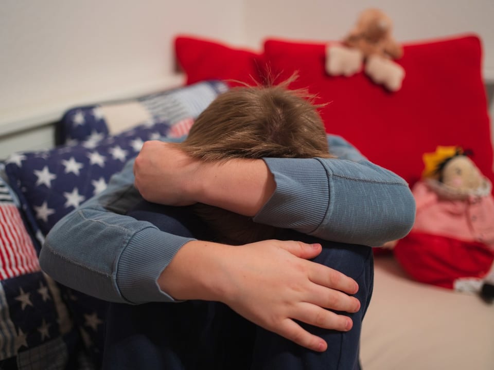 Kind sitzt zusammengekauert mit dem Kopf auf den Knien auf dem Sofa.