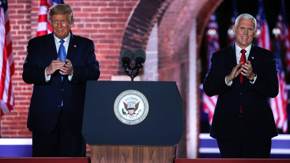 Zwei Männer hinter einem Rednerpult mit Mikrofonen und US-Flaggen im Hintergrund.