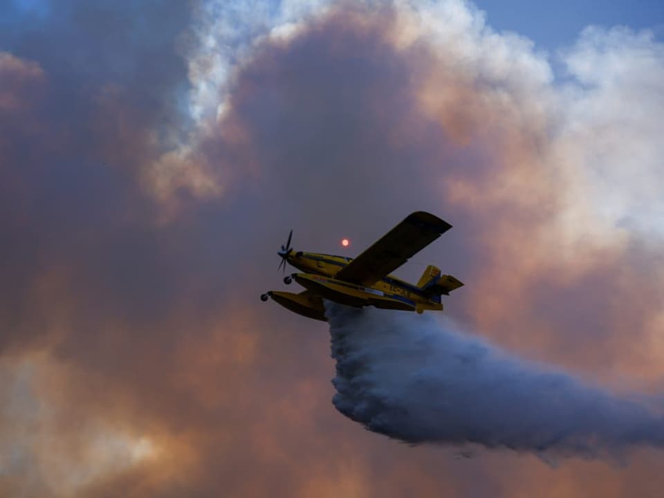 Gelbes Löschflugzeug fliegt durch Rauchwolken und sprüht Wasser.