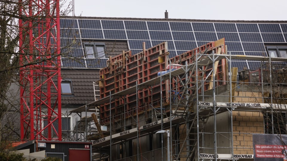 Bild von einer Baustelle, mit Krank und Baugerüsten im Vordergrund. Im Hintergrund ein Hausdach mit Sonnenkollektoren.