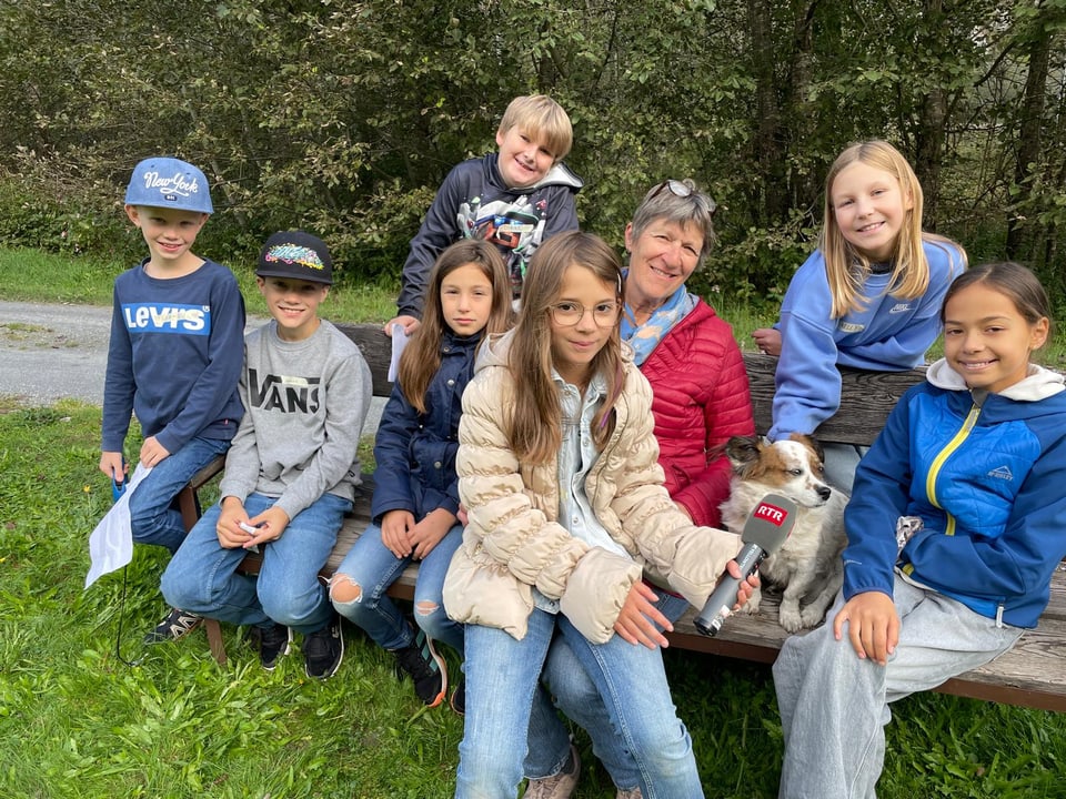 Kinder und eine Frau mit einem Mikrofon und einem Hund sitzen auf einer Bank draussen.