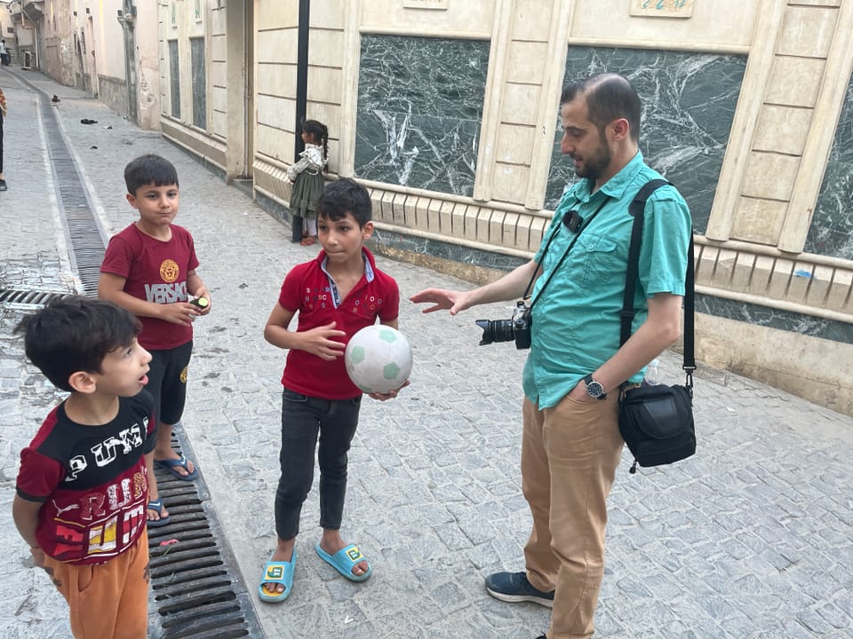 Erwachsener spricht mit drei Jungen, einer hält einen Fussball, auf einer gepflasterten Strasse.