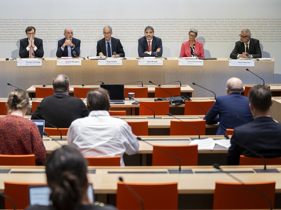 Sechs Personen sitzen bei einer Pressekonferenz an einem Tisch.