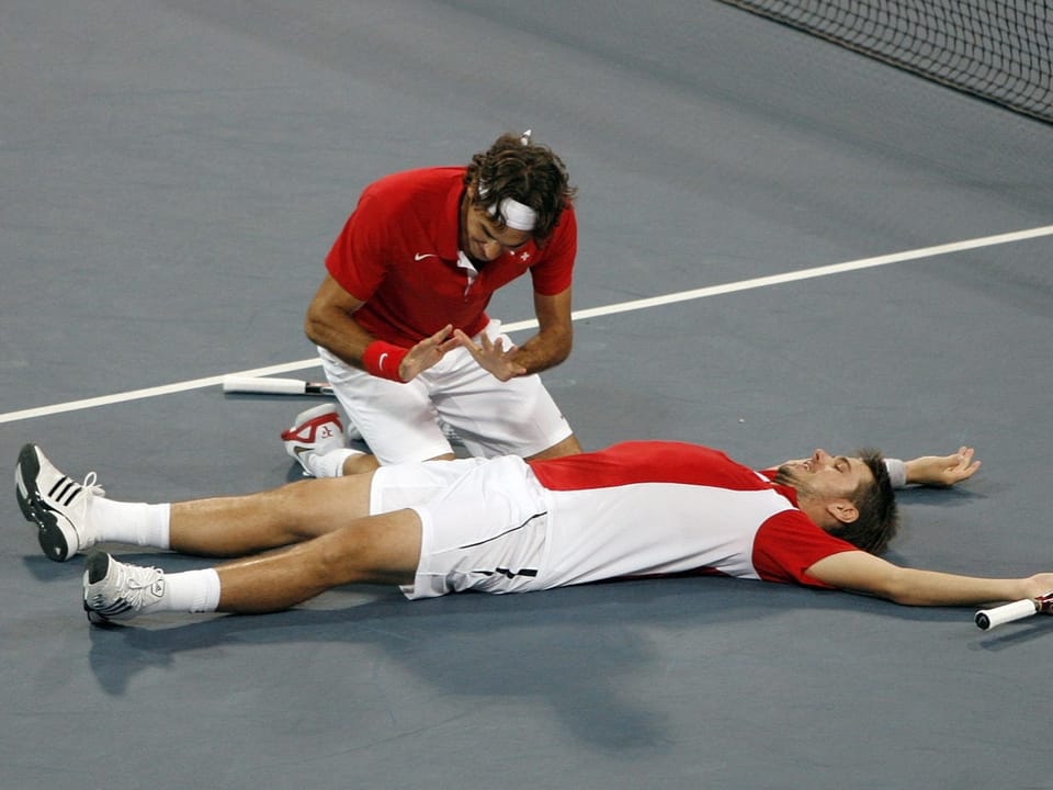 Zwei Tennisspieler feiern auf dem Platz, einer liegt auf dem Boden.