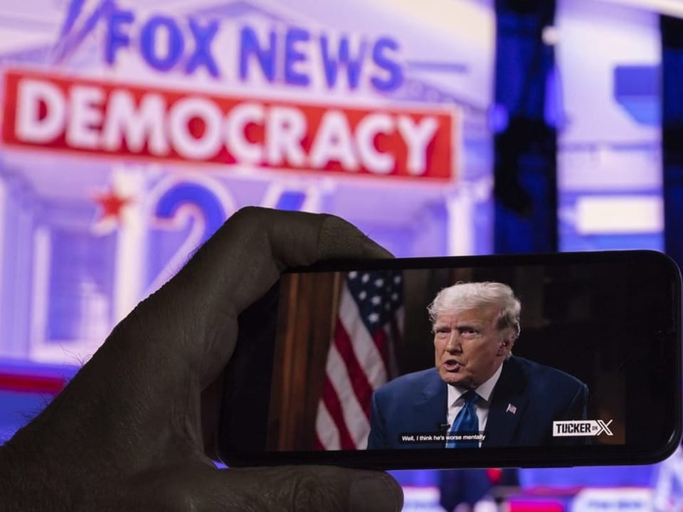 Hand hält Smartphone mit Trump-Interview vor Fox News Demokratie 24 Bildschirm.