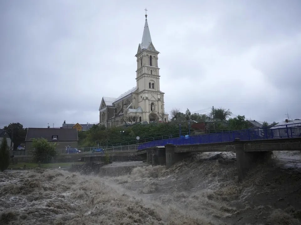 Kirche auf einem Hügel hinter einer Brücke und einem reissenden Fluss.