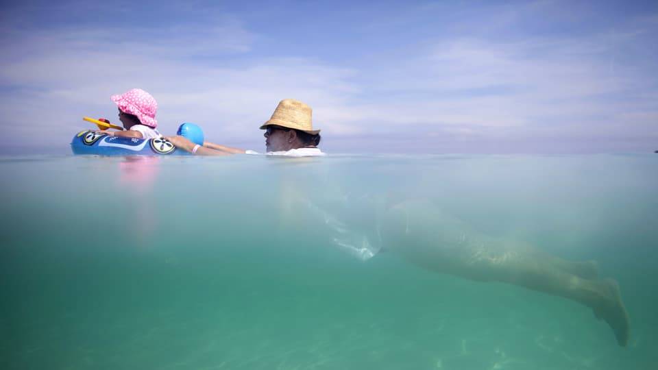 Frau mit Hut schwimmt im Meer und schiebt ein Baby im Schwimmring vor sich her.