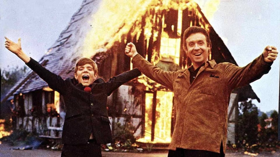 Peter Alexander und Heintje stehen jubelnd vor brennendem Gebäude.