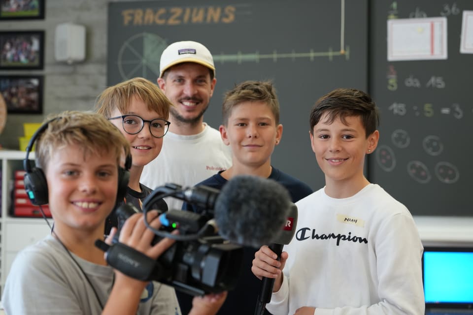 Fünf Jungen in einem Klassenzimmer, einer mit einer Kamera, fröhlich vor einer Tafel.