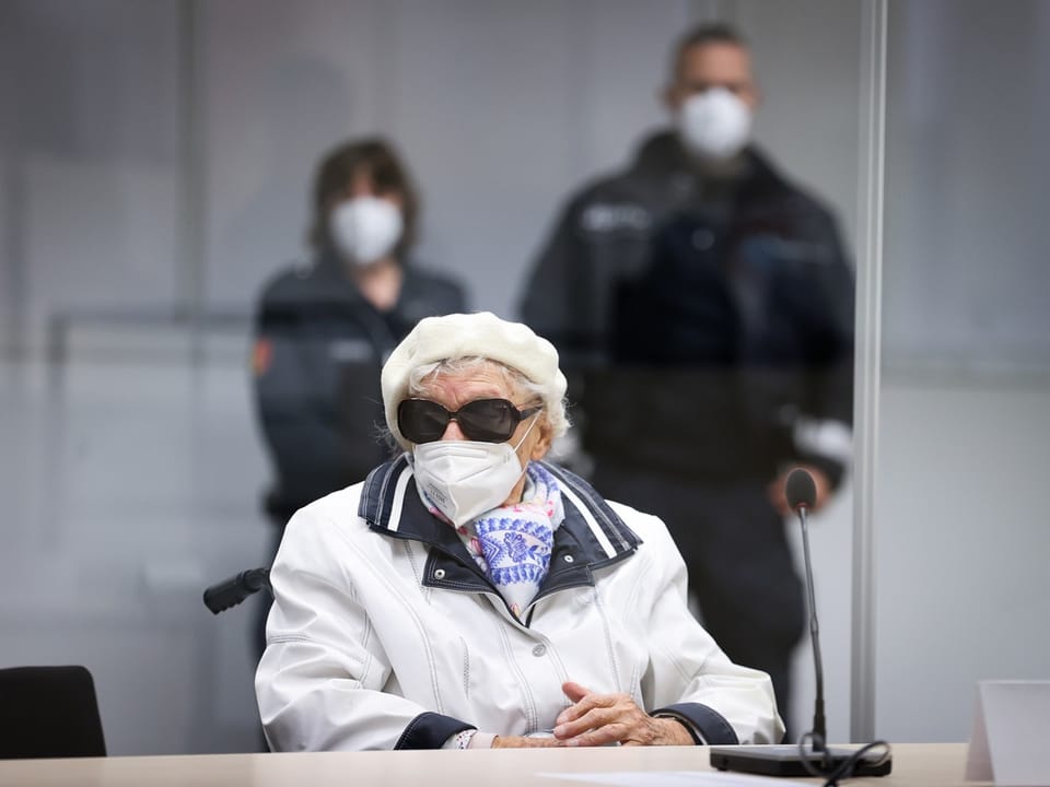 Ältere Frau mit Sonnenbrille und Maske sitzt vor einer Plexiglasscheibe.