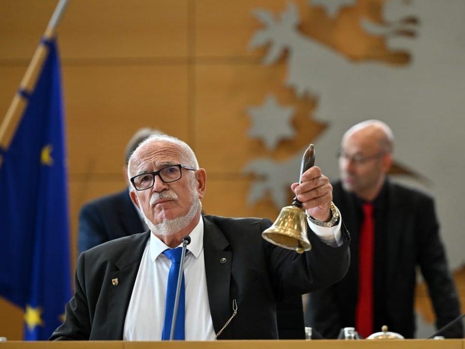 Älterer Mann im Anzug hält eine Glocke in einem Sitzungssaal.