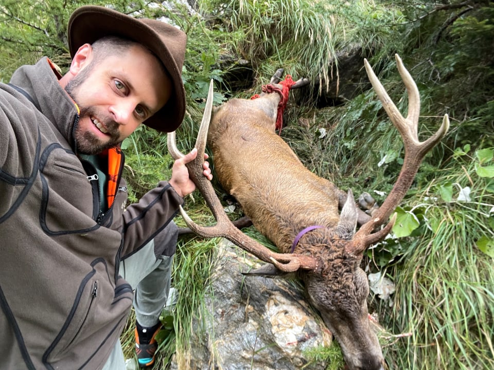 Mann mit Hut posiert neben erlegtem Hirsch im Wald.