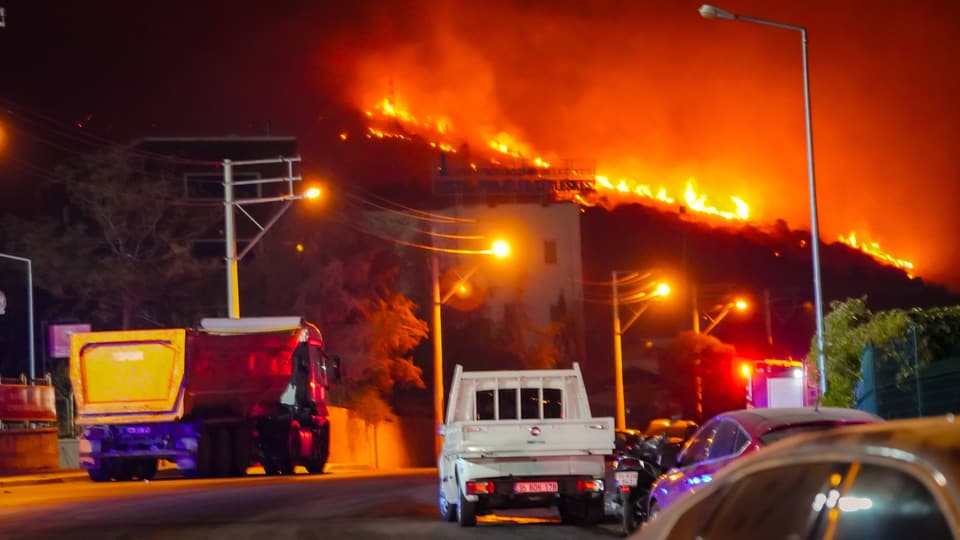 Nachtansicht eines Waldbrands am Hügel mit Fahrzeugen auf der Strasse.