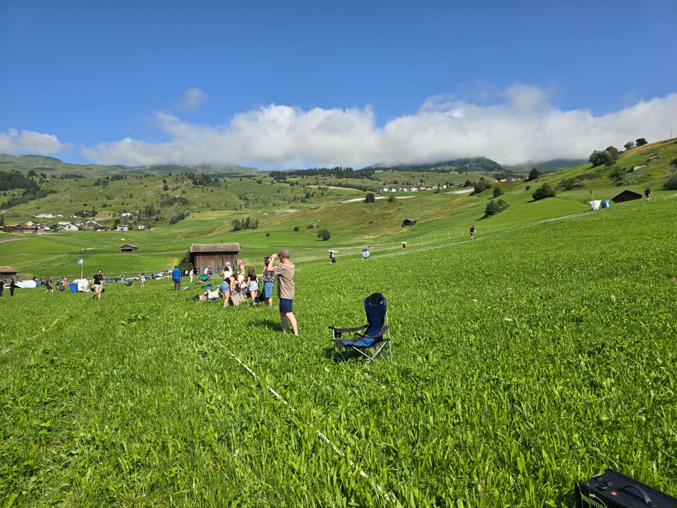 Leute auf grüner Wiese in Berglandschaft bei sonnigem Himmel.