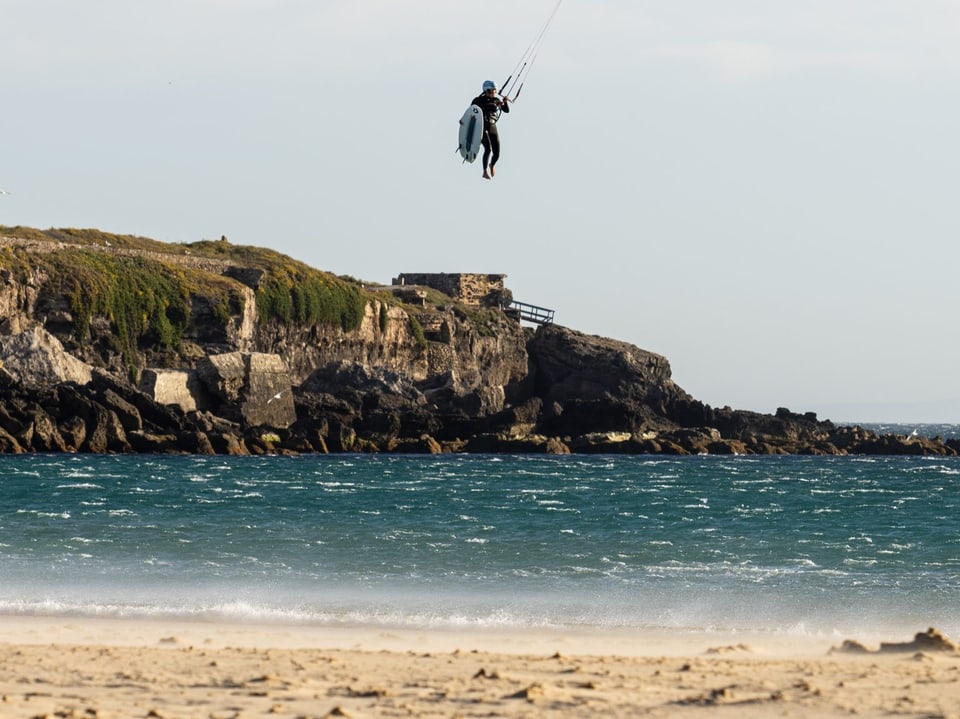 Kitesurfer schwebt über dem Meer vor felsiger Küste.