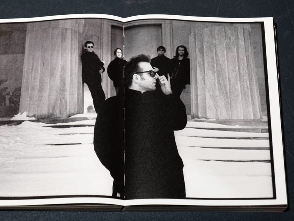 Ein Mann mit schwarzem Mantel und einer Sonnenbrille steht im Vordergrund, vier Männer im Hintergrund schauen ihm zu.t 