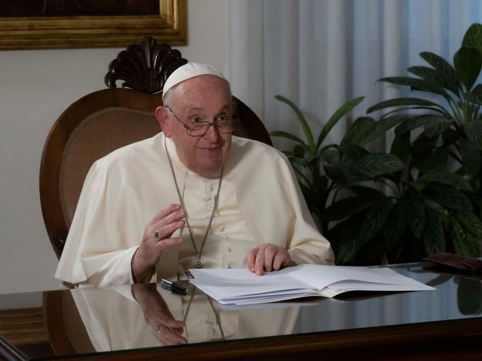 Papst Franziskus sitzt an einem Bürotisch, blättert in seinem Unterlagen und spricht zu Kamera