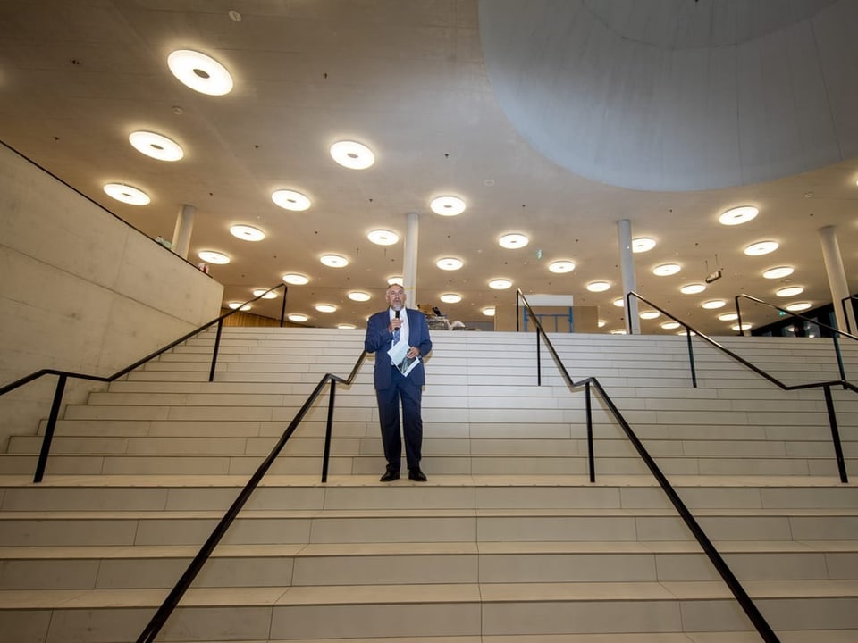 Mann steht ganz alleine auf grosser Treppe und hält ein Mikrophon vor sich.