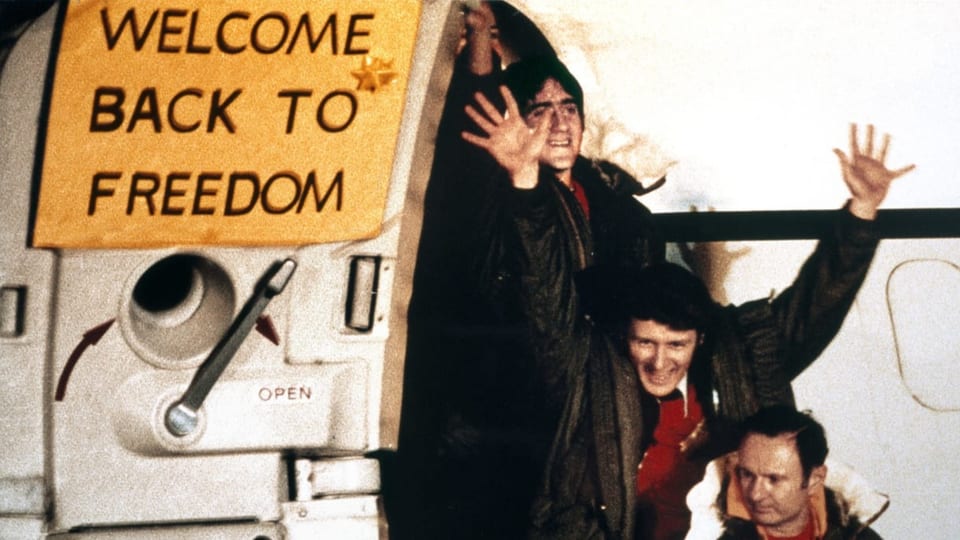 Drei Männer in schwarzen Jacken und roten Pullis steigen aus einem weissen Flugzeug aus. Links ein gelbes Schild.