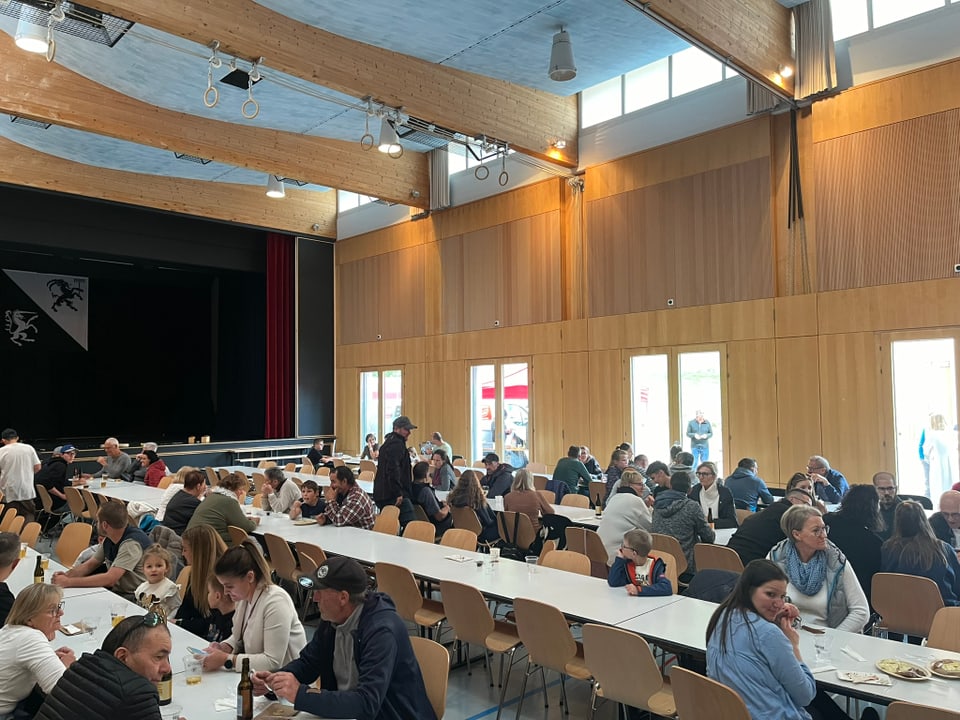 Menschen sitzen an Tischen in einer hellen, holzgetäfelten Halle.