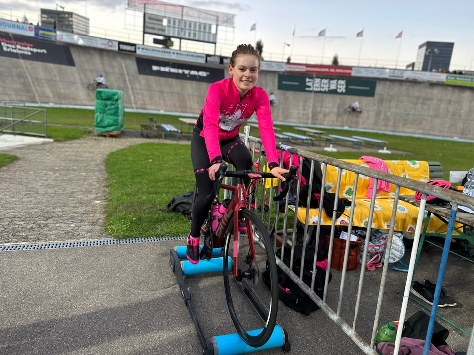 Ein Mädchen sitzt in pinkem Tenue auf einem Rennrad. Dieses steht auf drei Rollen. 