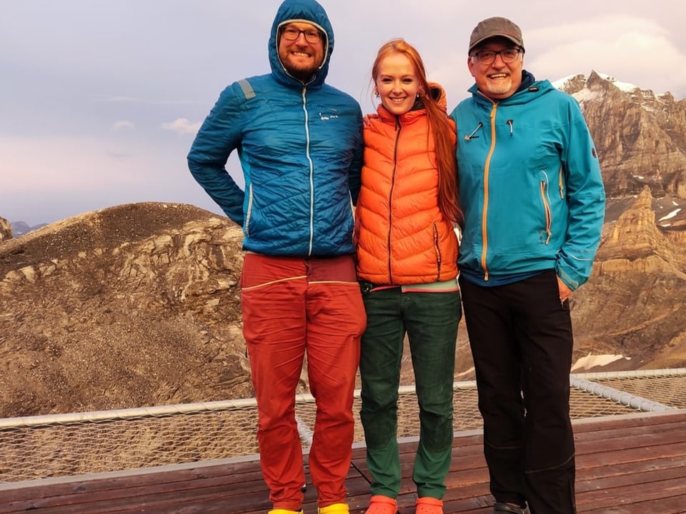 Drei Menschen im Vordergrund in Alpinkleidung. Im Hintergrund ein Bergmassiv.