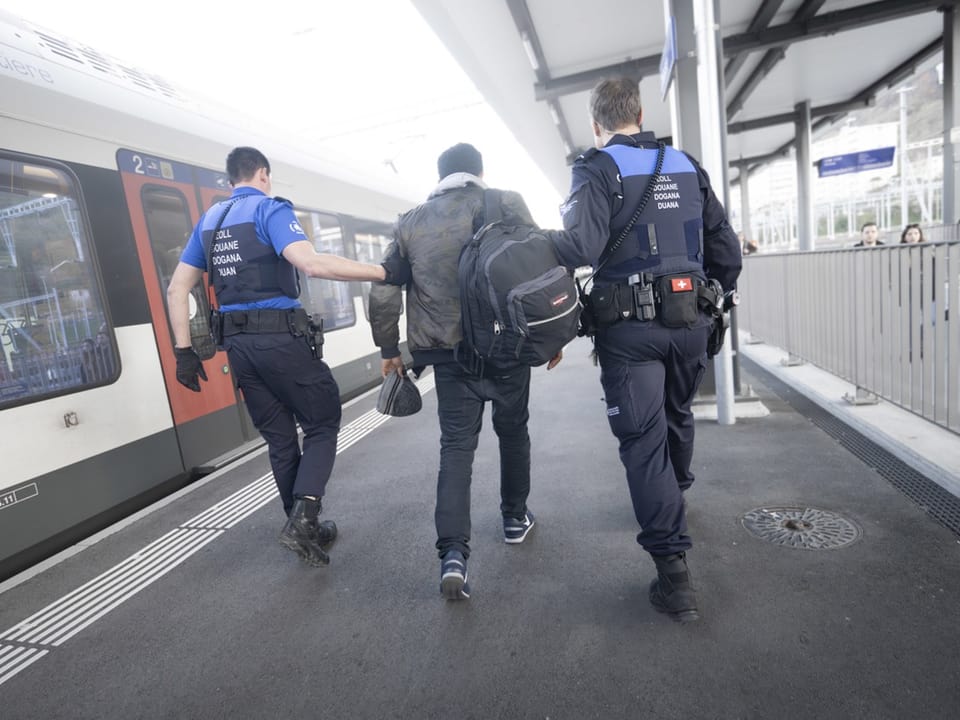 Zwei Zöllner begleiten eine Person auf einem Bahnsteig.