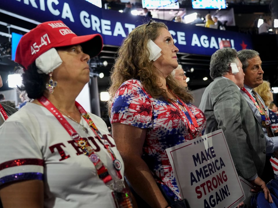 Personen mit Wattebäuschchen in den Ohren bei einer politischen Veranstaltung, Plakat mit 'Make America Strong Again!'