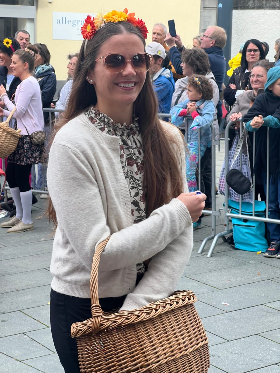 Frau mit Blumenkopfschmuck und Sonnenbrille bei einer Parade, hält einen Korb.