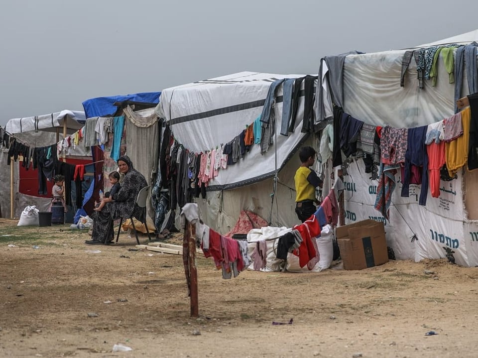 Flüchtlingslager mit Zelten und aufgehängter Wäsche, Menschen sitzend und stehend.