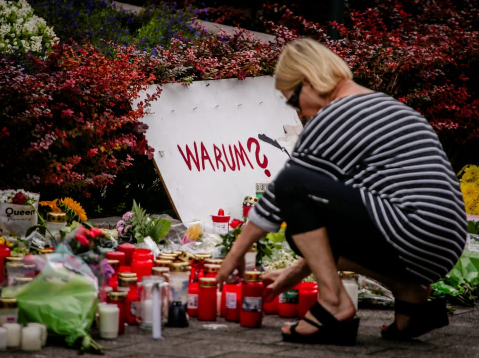 Frau kniet vor einem Gedenkort mit Kerzen und Blumen, Schild mit Aufschrift 'WARUM?'