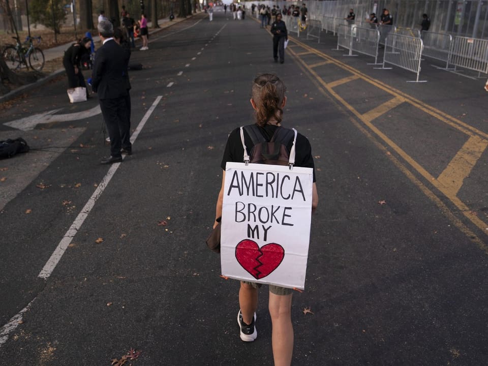 Person mit Schild 'America broke my heart' auf Strasse.