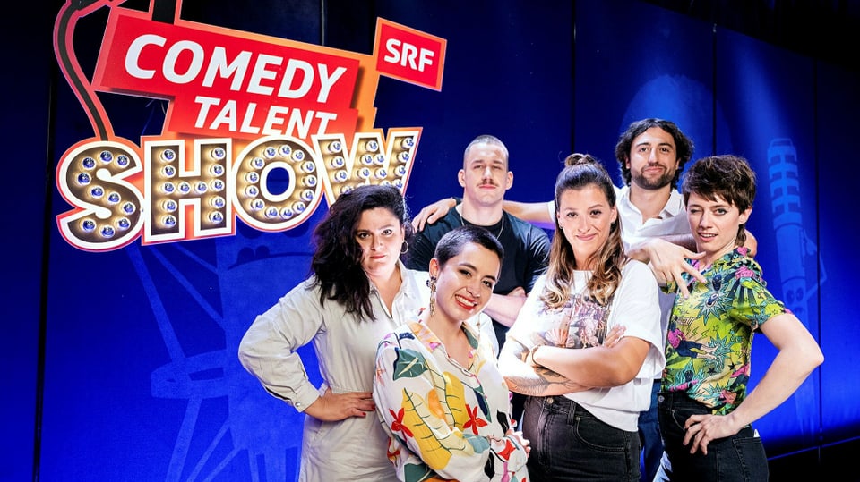 Eine Gruppe von Comedians posieren auf einer Bühne vor einem blauen Hintergrund und dem SRF Comedy Talent Show Logo.